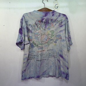 以下が含まれることがあります： 紫と青のタイダイ染めのTシャツ。Tシャツには、グレイトフル・デッドのバンドのグラフィックと「Grateful Dead」と「Don't miss every day special guest String Cheese Incident」の文字が書かれています。