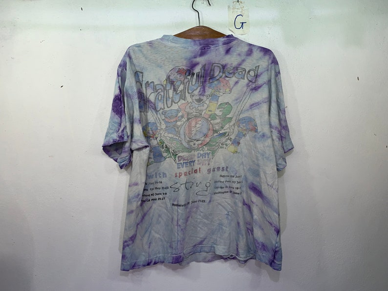 以下が含まれることがあります： 紫と青のタイダイ染めのTシャツ。Tシャツには、グレイトフル・デッドのバンドのグラフィックと「Grateful Dead」と「Don't miss every day special guest String Cheese Incident」の文字が書かれています。