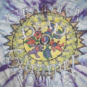 以下が含まれることがあります： タイダイ染めのTシャツに、銀色のグリッターで太陽と「Grateful Dead Summer Tour '93」の文字、そして踊るクマがデザインされています。