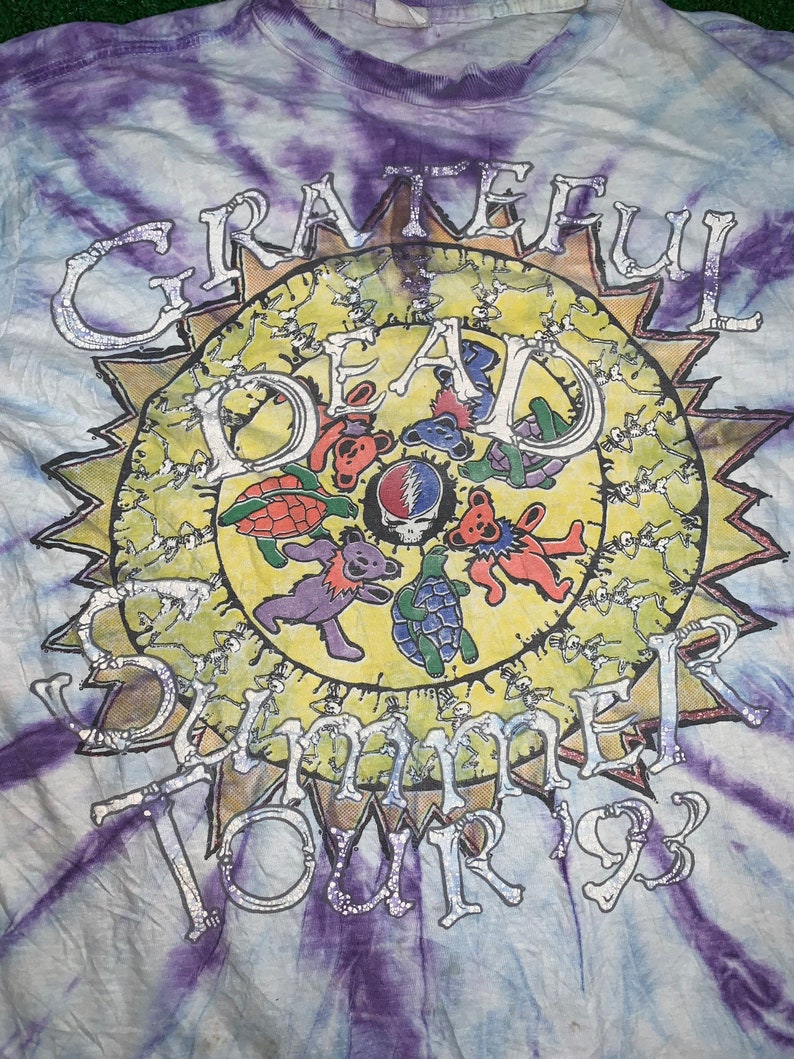 以下が含まれることがあります： タイダイ染めのTシャツに、銀色のグリッターで太陽と「Grateful Dead Summer Tour '93」の文字、そして踊るクマがデザインされています。