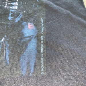 May include: Black t-shirt with a faded image of a person's face. The text "&copy; 2000 MARILYN MANSON UNDER LICENSE TO GIANT" is printed on the right side of the shirt.