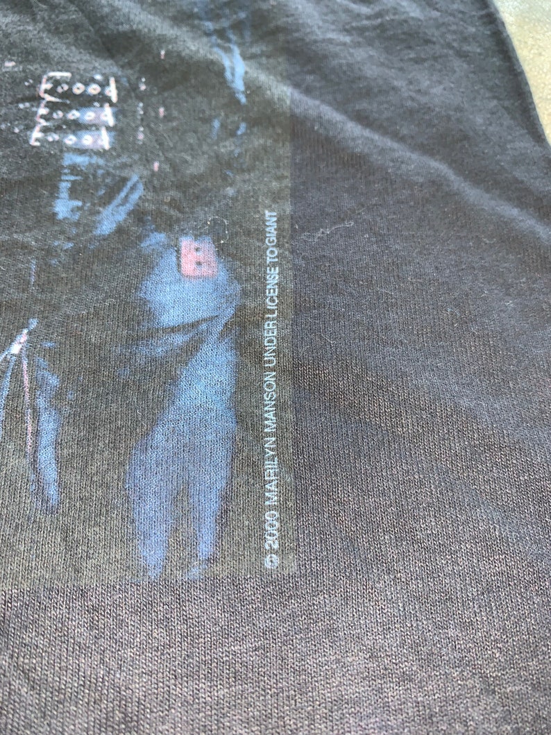 May include: Black t-shirt with a faded image of a person's face. The text "&copy; 2000 MARILYN MANSON UNDER LICENSE TO GIANT" is printed on the right side of the shirt.