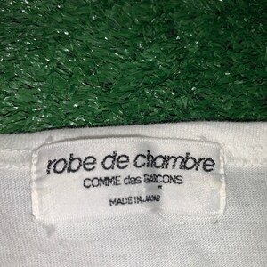 Pu&ograve; includere: Etichetta di abbigliamento bianca con testo nero che recita "robe de chambre COMME des GARCONS MADE IN JAPAN"