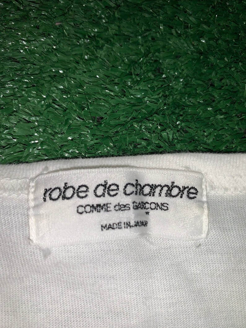 Pu&ograve; includere: Etichetta di abbigliamento bianca con testo nero che recita "robe de chambre COMME des GARCONS MADE IN JAPAN"
