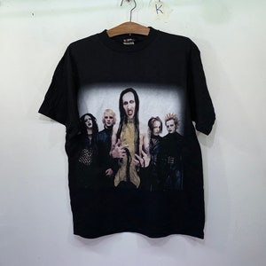 May include: Black t-shirt with a faded image of a band, featuring five people with long hair and dark clothing. The band members are standing in a row, with the person in the center wearing a gold shirt. The text "Marilyn Manson" is visible on the shirt.