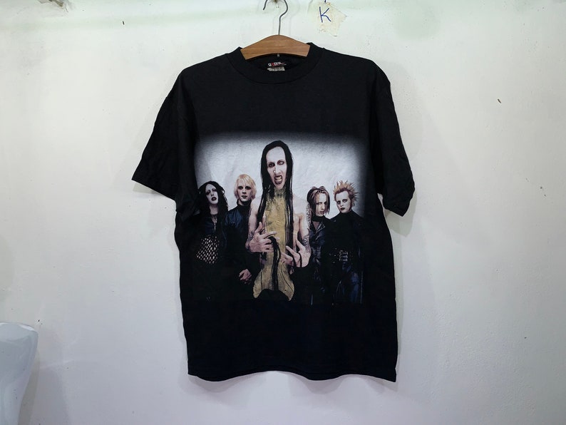 May include: Black t-shirt with a faded image of a band, featuring five people with long hair and dark clothing. The band members are standing in a row, with the person in the center wearing a gold shirt. The text "Marilyn Manson" is visible on the shirt.
