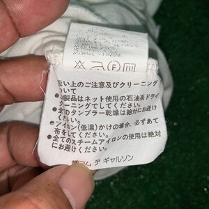 Pu&ograve; includere: Un'etichetta di cura bianca con testo giapponese e inglese. Il testo inglese dice: "JAVEL IN FR.  RECHAGE A MAL.  REPASSER A BASSE TEMPERATURE AVEC UNE PATTE.  NETTOYAGE A SEC.  SECHAGE A L'OMBRE." Il testo giapponese dice: "吸い上のご注意及びクリーニングについて" e "製品はネット使用の石油系ドライクリーニングでしてください。" e "全のタンブラー乾燥は絶対にお避けください。" e "アイロン(低温)かけの場合、必ずあて布をしてください。" e "全てのスチームアイロンの使用は絶対にお避けください。" e "(株)コムデギャルソン"