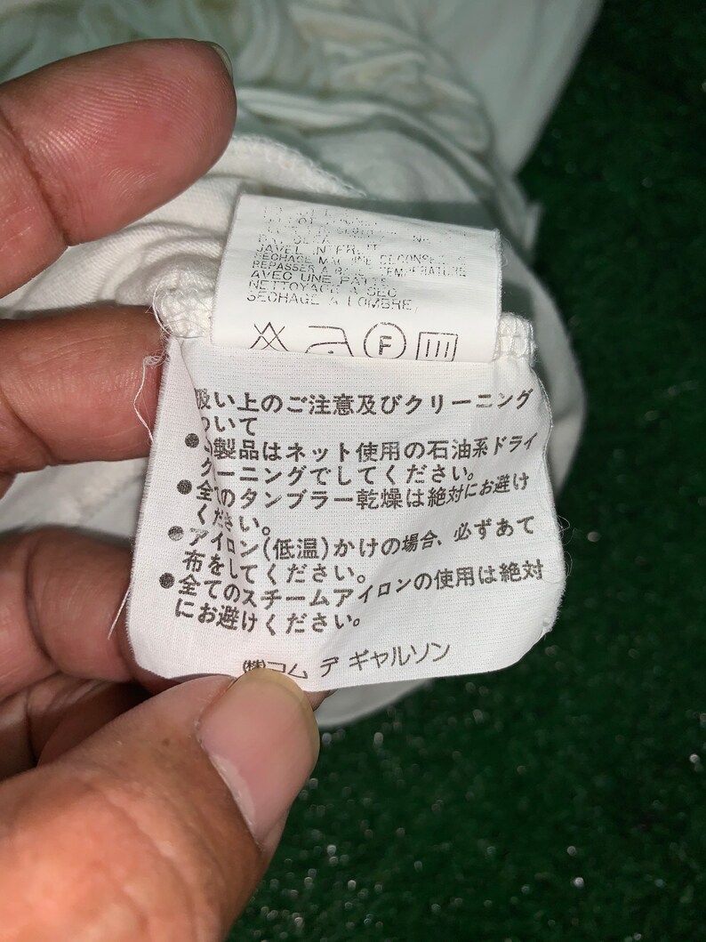 Pu&ograve; includere: Un'etichetta di cura bianca con testo giapponese e inglese. Il testo inglese dice: "JAVEL IN FR.  RECHAGE A MAL.  REPASSER A BASSE TEMPERATURE AVEC UNE PATTE.  NETTOYAGE A SEC.  SECHAGE A L'OMBRE." Il testo giapponese dice: "吸い上のご注意及びクリーニングについて" e "製品はネット使用の石油系ドライクリーニングでしてください。" e "全のタンブラー乾燥は絶対にお避けください。" e "アイロン(低温)かけの場合、必ずあて布をしてください。" e "全てのスチームアイロンの使用は絶対にお避けください。" e "(株)コムデギャルソン"