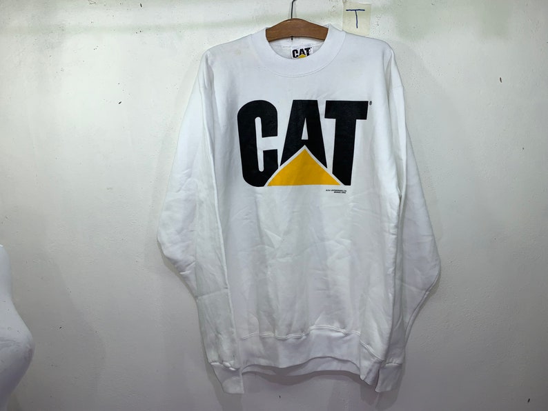 Pu&ograve; includere: Felpa bianca a maniche lunghe con un logo CAT nero e giallo. Il logo &egrave; un carattere grande e in grassetto con un triangolo giallo al centro.