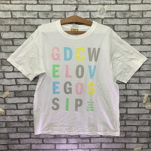 Puede incluir: Camiseta blanca con un estampado gráfico colorido que dice "GDCW ELOV EGOS SIP". El gráfico es de estilo bloque con cada letra de un color diferente.