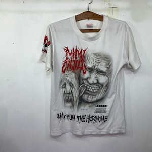 Puede incluir: Camiseta blanca con un estampado gráfico en blanco y negro de una cara distorsionada con el texto "M.I.A. " y "MAXIMUM THE HORMONE" en rojo.