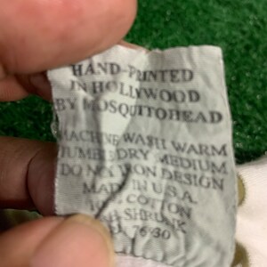 May include: White tag with black text that reads "HAND-PRINTED IN HOLLYWOOD BY MOSQUITOREAD MACHINE WASH WARM TUMBLE DRY MEDIUM DO NOT IRON DESIGN MADE IN USA COTTON SIZE UNK 7630"