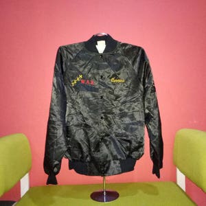 Może przedstawiać: Czarna satynowa kurtka bomberowa z haftowanym żółtym tekstem "Team War" i "Corvino" z przodu.