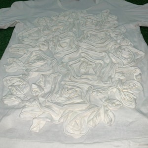 Pu&ograve; includere: Maglietta bianca con un grande disegno floreale 3D in tessuto bianco. Il disegno &egrave; centrato sulla parte anteriore della maglietta e assomiglia a un gruppo di rose.