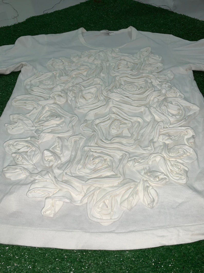 Pu&ograve; includere: Maglietta bianca con un grande disegno floreale 3D in tessuto bianco. Il disegno &egrave; centrato sulla parte anteriore della maglietta e assomiglia a un gruppo di rose.