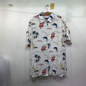 May include: White short-sleeved button-down shirt with a tropical print featuring palm trees, sailboats, and fish. The shirt has a blue, red, and green color scheme.