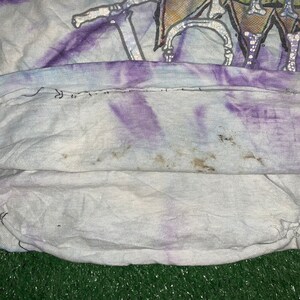 以下が含まれることがあります： 白地に紫と青のぼかし染めが施されたTシャツ。くたびれたような風合いがあり、柔らかく着心地の良い生地で作られています。