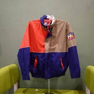 May include: A blue, red, and beige lightweight jacket with a zipper closure and a logo patch that says "HA" on the right sleeve.