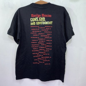 May include: Black t-shirt with a red and white graphic print that reads "Marilyn Manson Guns, God, and Government". The graphic also lists a tour schedule with cities in the United States, Canada, and Japan.