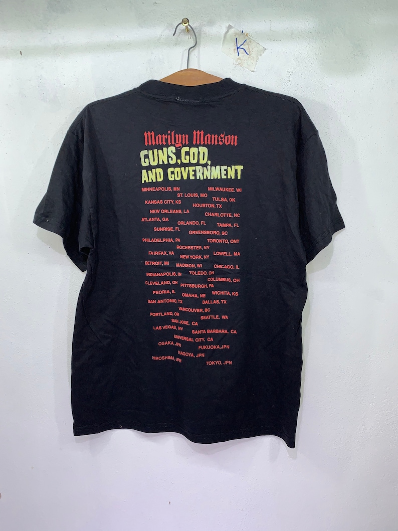 May include: Black t-shirt with a red and white graphic print that reads "Marilyn Manson Guns, God, and Government". The graphic also lists a tour schedule with cities in the United States, Canada, and Japan.