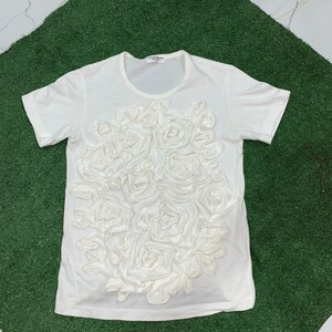 Pu&ograve; includere: Maglietta bianca con un grande disegno di rose bianche in 3D sul davanti. Le rose sono fatte di tessuto e sono disposte in un grappolo.