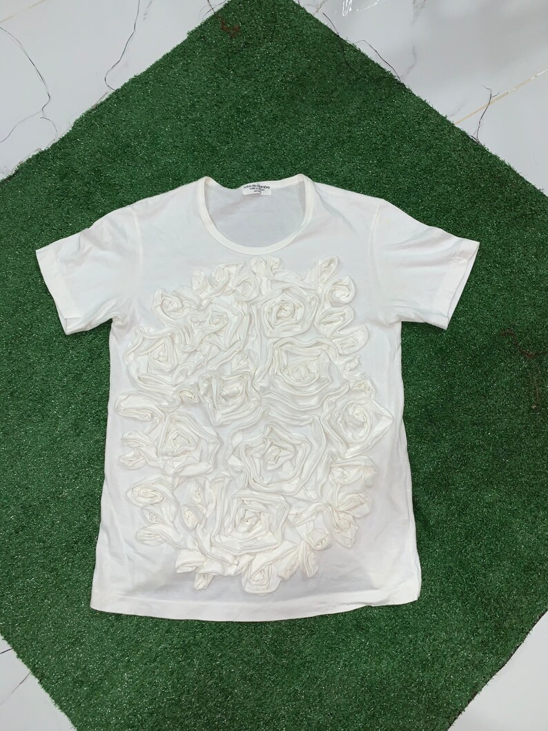 Pu&ograve; includere: Maglietta bianca con un grande disegno di rose bianche in 3D sul davanti. Le rose sono fatte di tessuto e sono disposte in un grappolo.