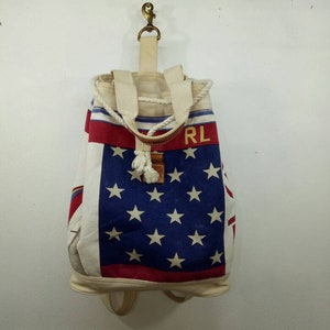 May include: A white canvas tote bag with a red, white, and blue American flag design. The bag has a brown leather handle and the letters "RL" are printed on the front.