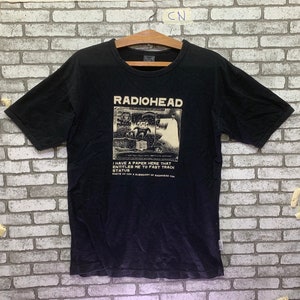 Può includere: Maglietta nera con una stampa bianca del gruppo Radiohead. La grafica include il nome del gruppo, un'immagine in bianco e nero e il testo "I have a paper here that entitles me to fast track status".