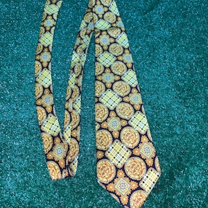 Puede incluir: Una corbata de seda con estampado negro y dorado con un diseño geométrico. La corbata tiene un extremo puntiagudo y un ancho estrecho.