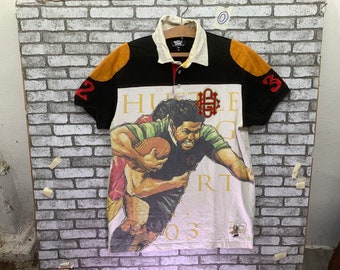 Può includere: Una maglia da rugby bianca, nera e arancione con una grafica di due giocatori in azione. La maglia ha il testo "Hustle Hard" e "03" stampato su di essa.