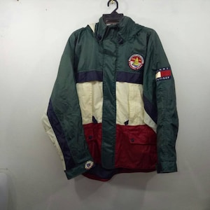May include: A green, white, red, and blue color block hooded jacket with a Tommy Hilfiger logo patch on the right sleeve. The jacket has a zipper closure and a drawstring hood.