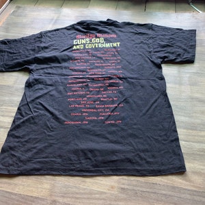 May include: Black t-shirt with a red and white graphic print of Marilyn Manson's Guns, God and Government tour dates. The tour dates include cities in the United States, Canada, and Japan.