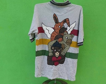 Può includere: Una maglietta grigia a maniche corte con un motivo a righe multicolore. La maglietta presenta una grafica di un coniglio marrone con le ali, un orso marrone e un orso nero che indossa un cappello blu. Gli orsi sono impilati uno sopra l'altro, con il coniglio in cima.