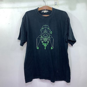 May include: Black t-shirt with a neon green graphic of a robot-like figure and the text "WHAT AM I?". The shirt is hanging on a wooden hanger against a white wall. The t-shirt is a casual wear item.