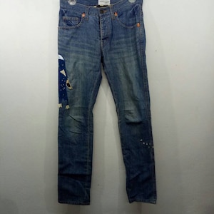 Puede incluir: Vaqueros de mezclilla azul con un diseño de patchwork en la pierna izquierda. Los vaqueros tienen cierre de botón y cinco bolsillos.