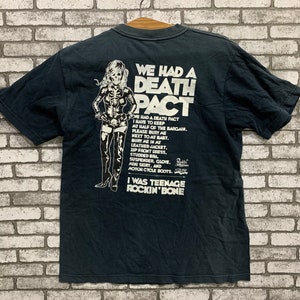 Può includere: Maglietta nera con una stampa grafica bianca di una donna in un outfit di pelle e il testo "We had a Death Pact". Il testo continua con un elenco di articoli, tra cui "giacca di pelle", "abito con zip frontale", "reggiseno con borchie", "bretelle", "guanto", "minigonna" e "stivali da motocicletta". Il testo termina con "I was teenage rockin' bone".