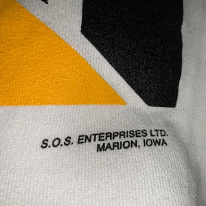 Pu&ograve; includere: Maglietta di cotone bianco con un design geometrico nero e giallo. Il testo "S.O.S. ENTERPRISES LTD. MARION, IOWA" &egrave; stampato sul fondo della maglietta.