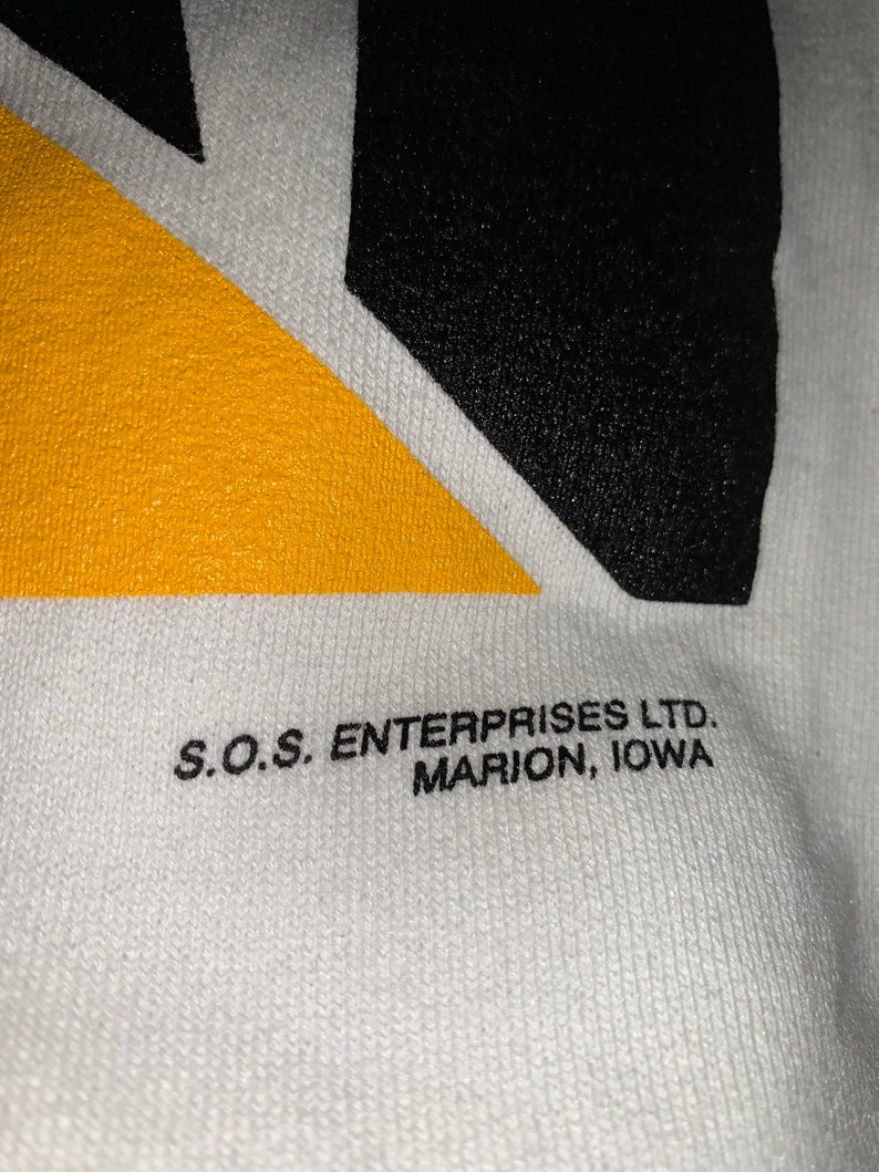 Pu&ograve; includere: Maglietta di cotone bianco con un design geometrico nero e giallo. Il testo "S.O.S. ENTERPRISES LTD. MARION, IOWA" &egrave; stampato sul fondo della maglietta.