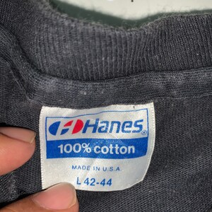 May include: A black Hanes t-shirt tag with white text that reads "100% cotton" and "Made in U.S.A." The tag also has a size label that reads "L 42-44".