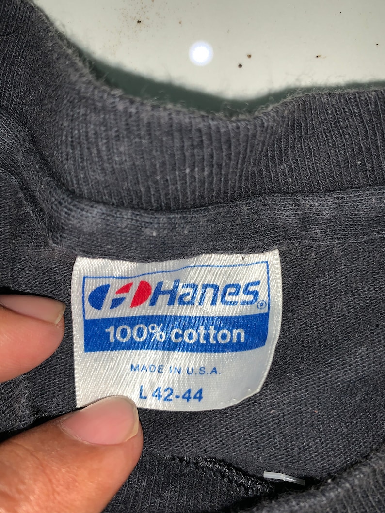 May include: A black Hanes t-shirt tag with white text that reads "100% cotton" and "Made in U.S.A." The tag also has a size label that reads "L 42-44".