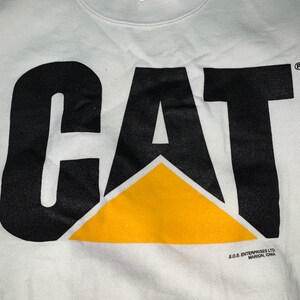 Pu&ograve; includere: Felpa bianca con un logo "CAT" nero con un triangolo giallo. Il logo &egrave; stampato sul davanti della felpa. La felpa ha un collo rotondo e maniche lunghe. Il testo "S.O.S. ENTERPRISES LTD. MARION, IOWA" &egrave; stampato sul fondo della felpa.