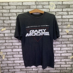 以下が含まれることがあります： 三角形の中に「Corridors of Power」と「Gary Moore」の文字が入った白いグラフィックプリントが施された黒いTシャツです。