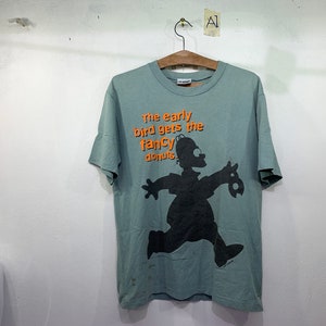 Könnte beinhalten: Ein hellblaues T-Shirt mit einer laufenden Comicfigur, die einen Donut hält. Der Text auf dem Shirt lautet "The early bird gets the fancy donuts."