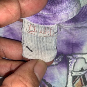 以下が含まれることがあります： 紫と白のタイダイ染めのシャツに、黒い文字で「CLBT」と書かれた白いタグのアップ。