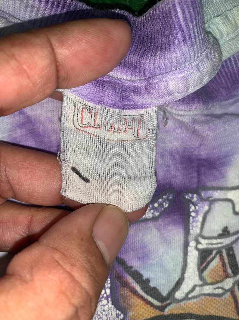 以下が含まれることがあります： 紫と白のタイダイ染めのシャツに、黒い文字で「CLBT」と書かれた白いタグのアップ。