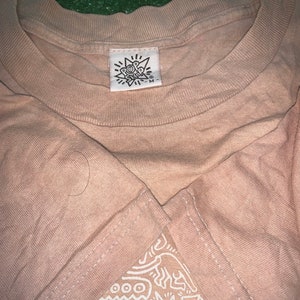 May include: A beige t-shirt with a white graphic print of abstract shapes and lines. The shirt has a tag that says "Pop Shop" with a star and the letter "M" on it.