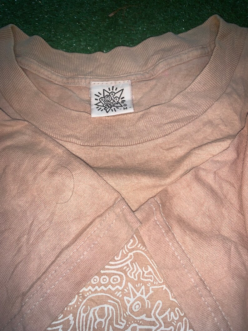 May include: A beige t-shirt with a white graphic print of abstract shapes and lines. The shirt has a tag that says "Pop Shop" with a star and the letter "M" on it.