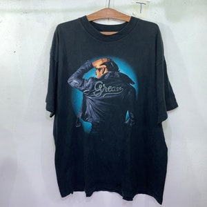 Può includere: T-shirt nero con una stampa grafica di un uomo con una giacca di pelle con la parola "Grease" sul retro.