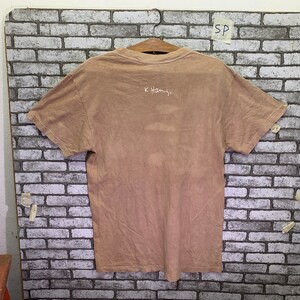 May include: A light brown t-shirt with a white inscription that reads "K.Hamj."
