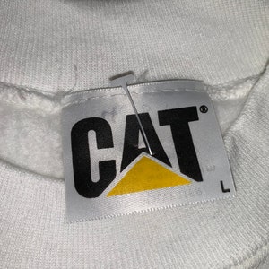 Pu&ograve; includere: Felpa bianca con un'etichetta logo CAT nera e gialla. L'etichetta ha la parola "CAT" in lettere nere con un triangolo giallo sotto. L'etichetta ha anche un'etichetta di taglia che dice "L".